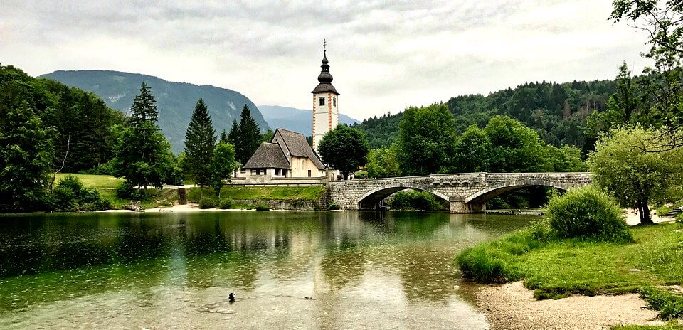 A church with a tall steeple next to a stone bridge and lake surrounded by lush green hills, Slovenia