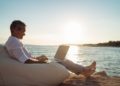 Senior man working on his laptop lying on deck chair on the beach during sunset