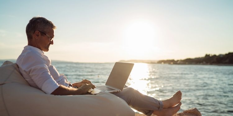 Senior man working on his laptop lying on deck chair on the beach during sunset