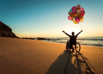 A person on a wheelchair with colorful balloons at the beach. disability friendly countries