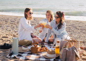 Three young beautiful happy women female friends having cozy summer picnic with lemonade, fresh bread and fruits on a beach