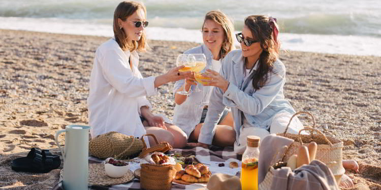 Three young beautiful happy women female friends having cozy summer picnic with lemonade, fresh bread and fruits on a beach