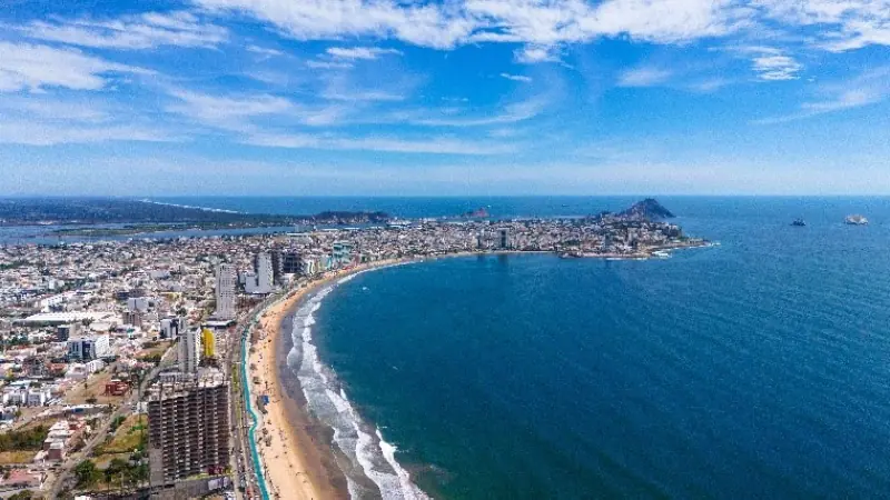 Aerial view of Mazatlán, Mexico, showing the curved coastline, sandy beach, and cityscape against a bright blue sky.