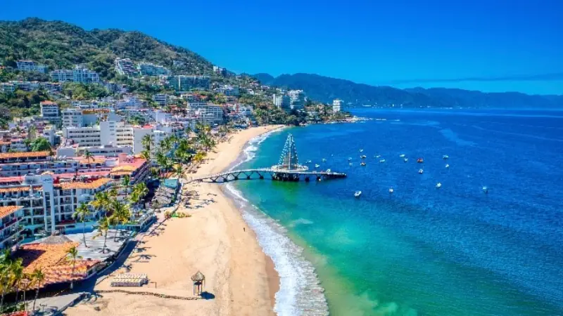 Scenic view of Puerto Vallarta, Mexico, with sandy beach, coastal buildings, and the Los Muertos Pier over turquoise water.