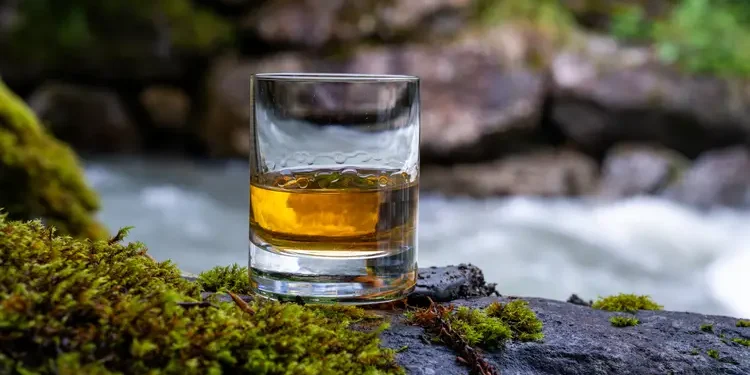 Glass of strong scotch single malt whisky with fast flowing mountain river on background. liquid gold