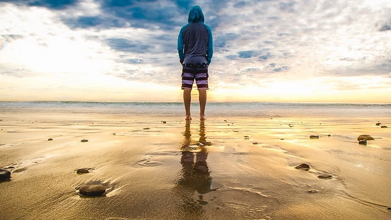 man in shorts and a hooded shirt standing on beach looking at the ocean