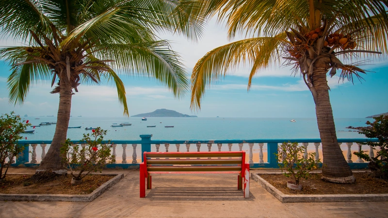 View of Taboga with palms surrounding a red bench with a view towards an island and a beach with boats. taboga