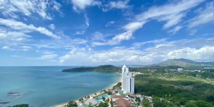 A coastal resort with a beachfront hotel, turquoise ocean views, and lush green hills under a bright blue sky.