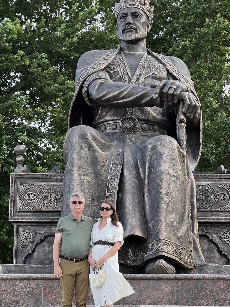 ief Simon and Kathleen Peddicord pose in front of the grand statue of Amir Timur in Samarkand, Uzbekistan. The bronze statue depicts the historic conqueror seated on an ornate throne, wearing a crown and intricately detailed royal robes, with one hand resting on a cane. Behind them, leafy green trees provide a natural backdrop on a sunny day.