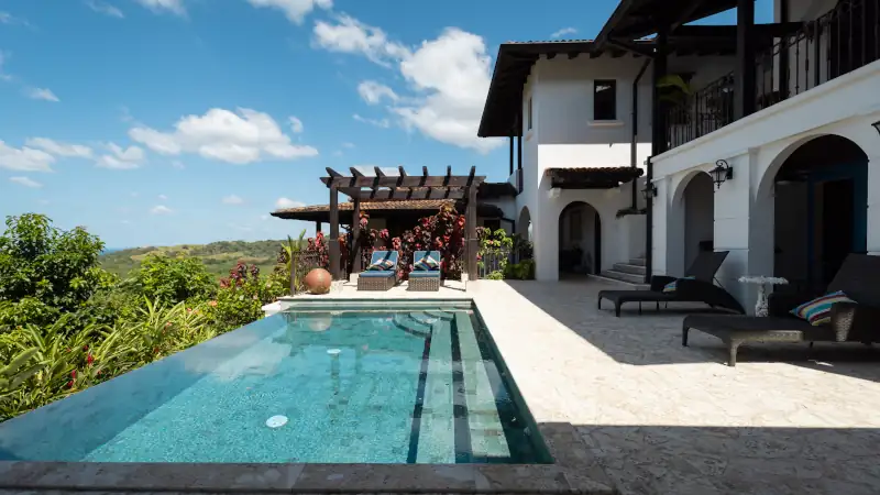 Luxury hillside villa in Los Islotes featuring an infinity pool, stone patio, lounge chairs, and pergola seating overlooking lush tropical greenery under a bright blue sky.