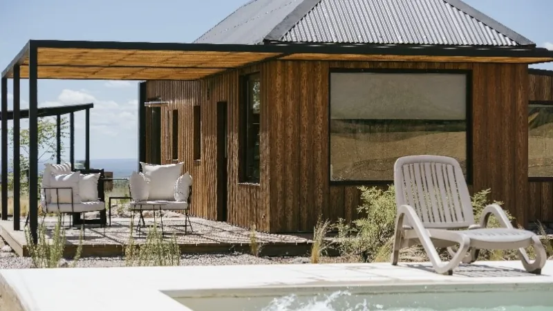 Modern wooden cabin with large windows and a shaded patio, featuring outdoor seating and a lounge chair beside a pool.