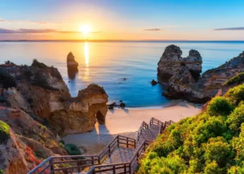 Sunrise over a rocky beach with cliffs and a wooden staircase leading down to the sand in Algarve, Portugal.