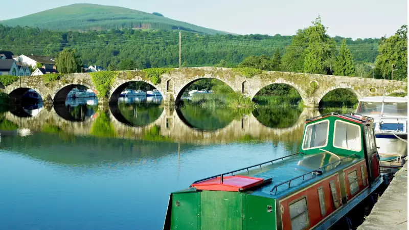 A canal boat docked near a stone arch bridge over calm water, with green hills and trees in the background.