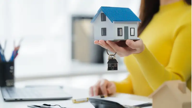 A woman presents a miniature house model, accompanied by keys, symbolizing a pivotal moment and a fresh start.