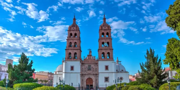Front view of Durango Cathedral with twin bell towers under a bright blue sky, surrounded by green trees and a plaza.