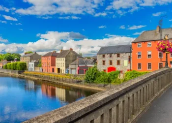 Colorful riverside buildings in Kilkenny, Ireland, viewed from a stone bridge under a blue sky.