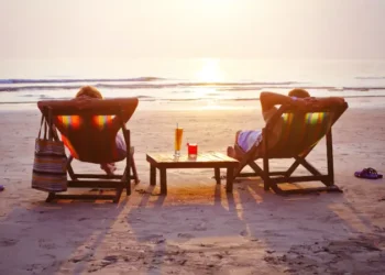 Two people relax in beach chairs facing the ocean at sunset, with drinks on a small table between them.