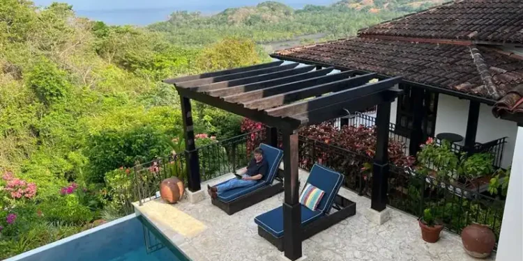 Tropical patio with lounge chairs, pool, lush greenery, and mountain view. One person relaxing.