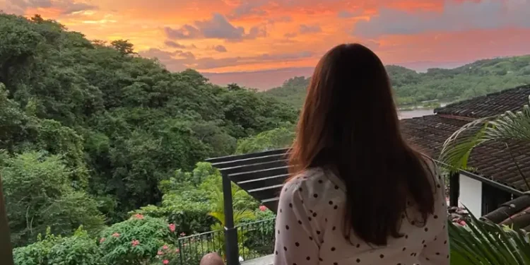 Kathleen Peddicord stands on a terrace, facing away from the camera, overlooking a lush green landscape and a vibrant orange-pink sunset sky.