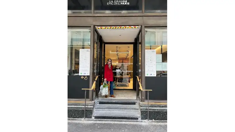 standing at the entrance of La Grande Épicerie de Paris, holding shopping bags on the steps outside the store.