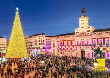 Festive crowd gathers around a great golden Christmas tree in the Puerta del Sol square, with the historic Real Casa de Correos Palace illuminated at dusk.