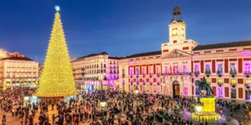Festive crowd gathers around a great golden Christmas tree in the Puerta del Sol square, with the historic Real Casa de Correos Palace illuminated at dusk.