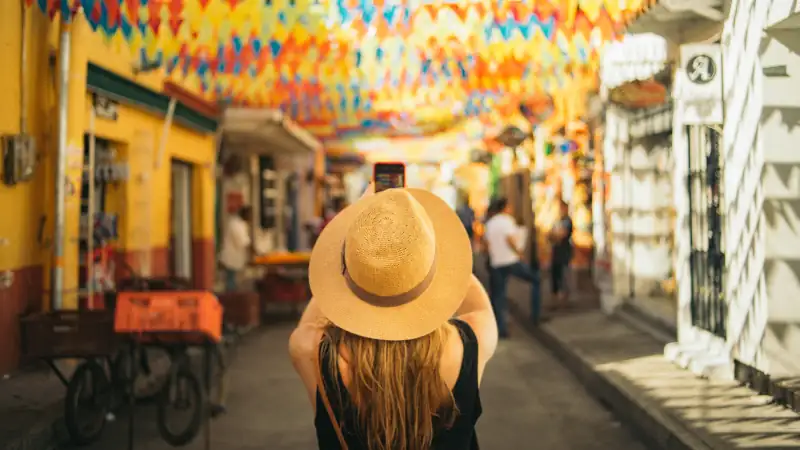 Colorful street scene with a woman wearing a straw hat taking a photo as vibrant flags hang overhead in a lively, sunlit neighborhood.