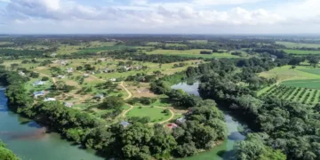 An aerial view of a lush rural area with a winding river, dense green trees, scattered homes, and farmland stretching into the distance under a partly cloudy sky.