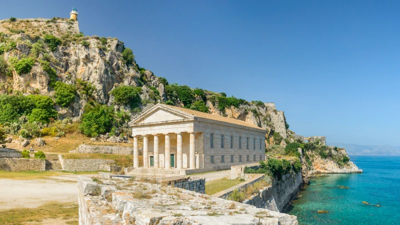 A side-angle view of the Church of St. George in the Old Fortress of Corfu, Greece, showcasing its Doric temple-style architecture on the seashore under a clear blue sky. 537813828