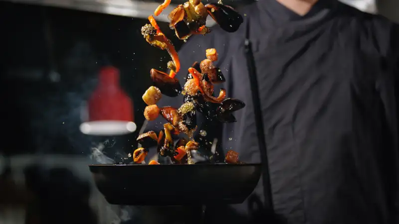 Chef tossing sauteed vegetables in pan over stove in kitchen, dark background