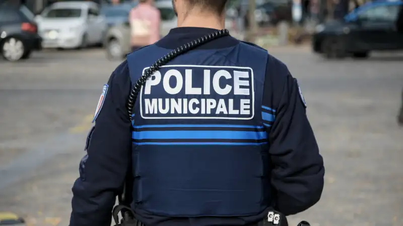 A municipal police officer seen from behind wearing a blue uniform and radio equipment while standing on a city street during daytime patrol.