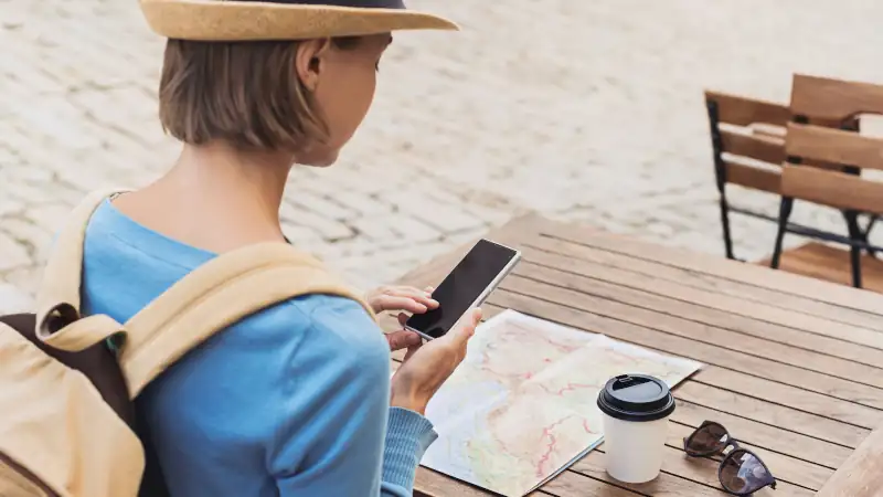 Young traveller woman sitting in a cafe terrace and planning trip with map and smartphone.