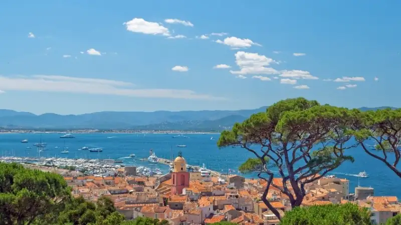 Panoramic coastal view of a Mediterranean seaside town with terracotta rooftops, a marina filled with yachts, turquoise waters, and pine trees overlooking the bay under a clear blue sky.