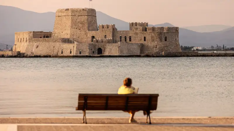 A solitary figure sits on a bench, gazing at the historic Bourtzi fortress in Nafplio, Greece
