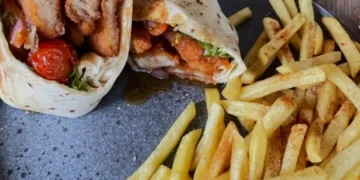 Crispy chicken wrap filled with fresh lettuce, pickled onions, roasted tomatoes, and sauce, served with seasoned French fries on a ceramic plate.