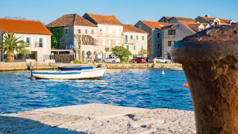 Mooring bollard in Tivat, Montenegro. A small white boat is anchored in the blue water. Colorful buildings line the shore under a clear sky.