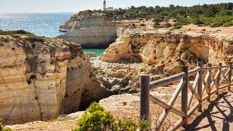 Scenic golden limestone cliffs overlooking turquoise waters, with a historic lighthouse perched on a rocky headland along the Portuguese coast.