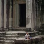 A child sits cross-legged outside a stone temple in Angkor Wat, Cambodia