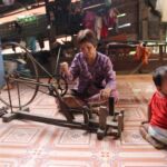 A Cambodian woman weaves traditional cloth, while her baby looks on.